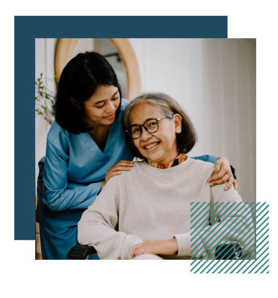 A woman is sitting in a wheelchair. She is smiling at the camera. Another woman in medical scrubs stands behind her and has her arms wrapped around the woman's shoulders.